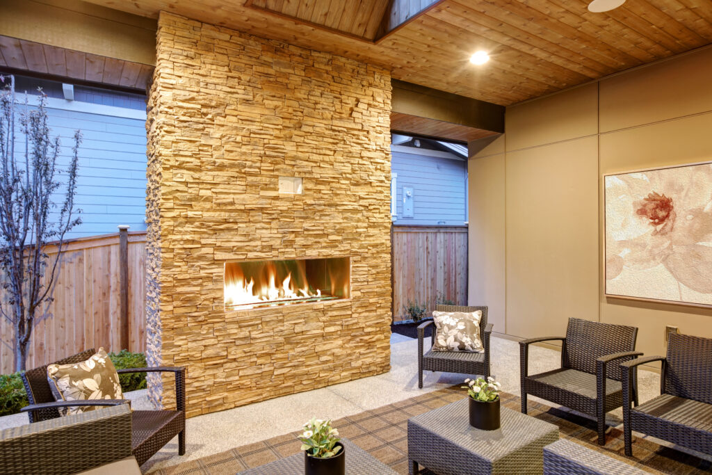 Dreamy outdoor covered patio with stone fireplace, a beadboard ceiling with skylight, rattan armchairs, sofa and ottomans overlooking the expansive backyard. Northwest,USA