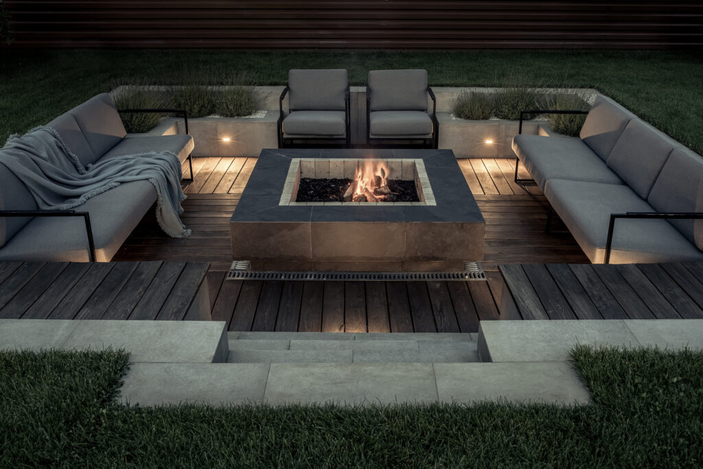 Zone for relax with a wooden floor and a tiled stair outdoors. There is a burning fire pit, gray sofas and armchairs, plaid, luminous lamps. Horizontal.