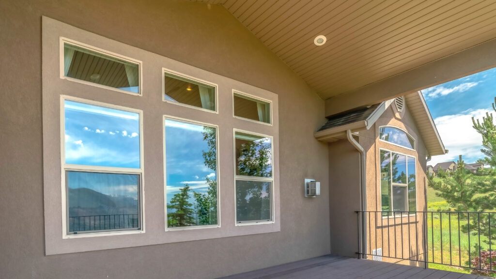 Panorama Sunlit deck of a home with wooden floor metal railing and white ceiling. The house has shiny windows that reflect a scenic view of mountain, trees, and sky.