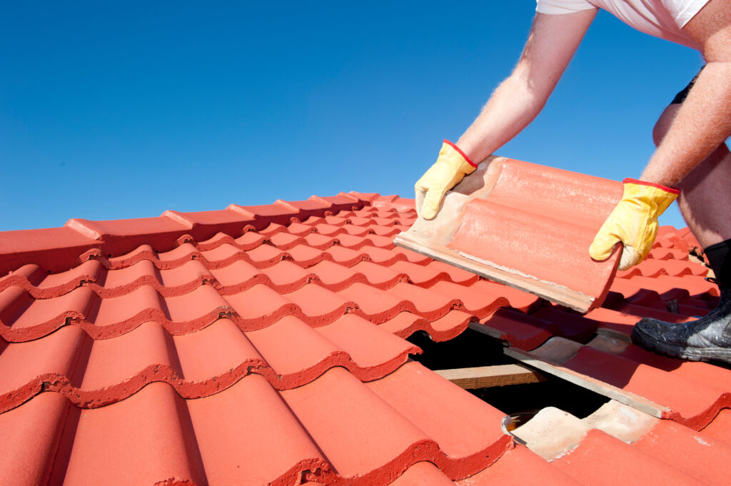 Roof repair, worker with yellow gloves replacing red tiles or shingles on house with blue sky as background and copy space.